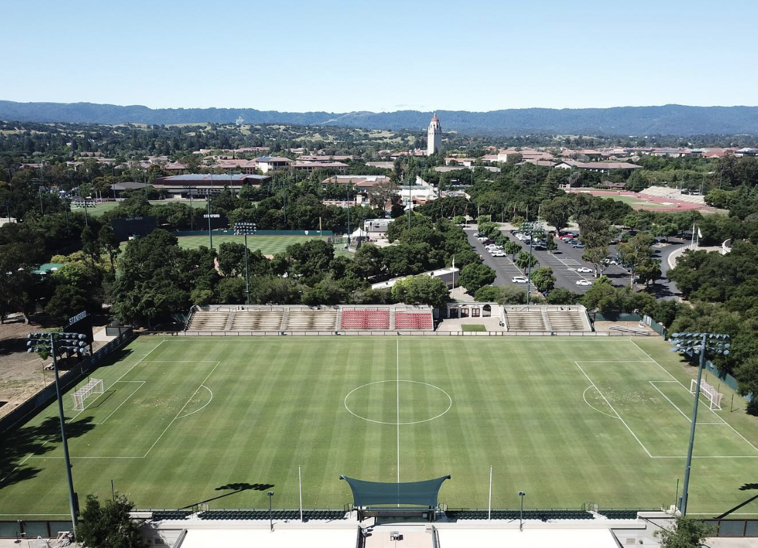 Aerial view of Stanford's Cagan Stadium soccer field, with red and white bleacher seating and Hoover Tower visible in the background.