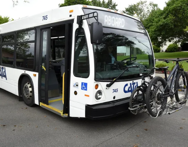 A white CATA transit bus, No. 745, parked with its front door open and a bicycle loaded onto the front bike rack, trees and a building visible in the background.