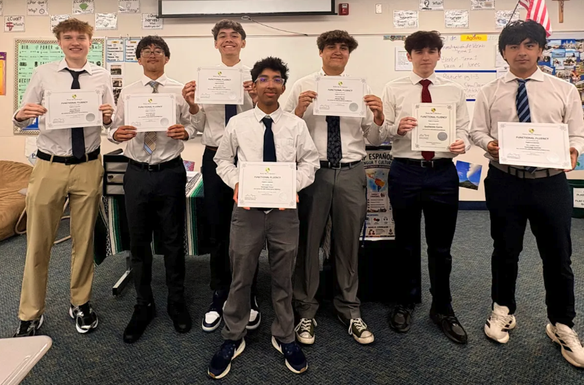 Seven Detroit Catholic Central High School students in dress shirts and ties stand in a classroom holding Global Seal of Biliteracy certificates.