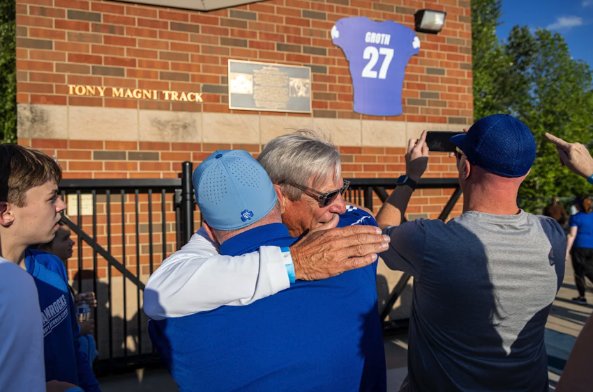 Two men embrace outside Catholic Central's stadium beneath a banner marking the retired No. 27 jersey, near a sign reading "Tony Magni Track."