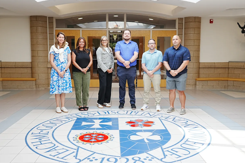 Six new Catholic Central High School staff members stand together in the school lobby atop the school's crest floor mosaic reading "Teach Me Goodness, Discipline, and Knowledge, 1928."