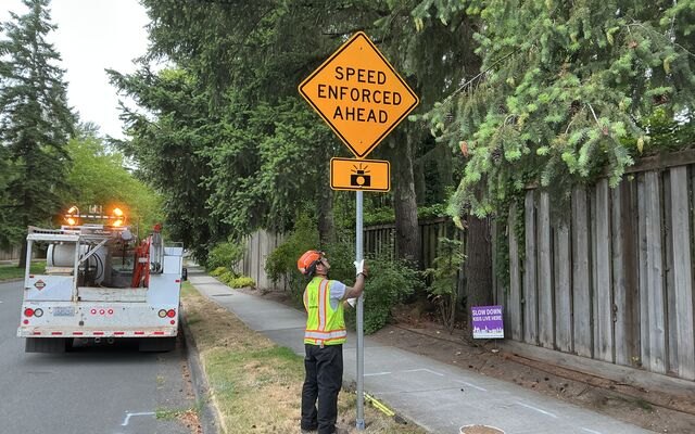 Worker in safety vest installs a yellow Speed Enforced Ahead sign on a pole.