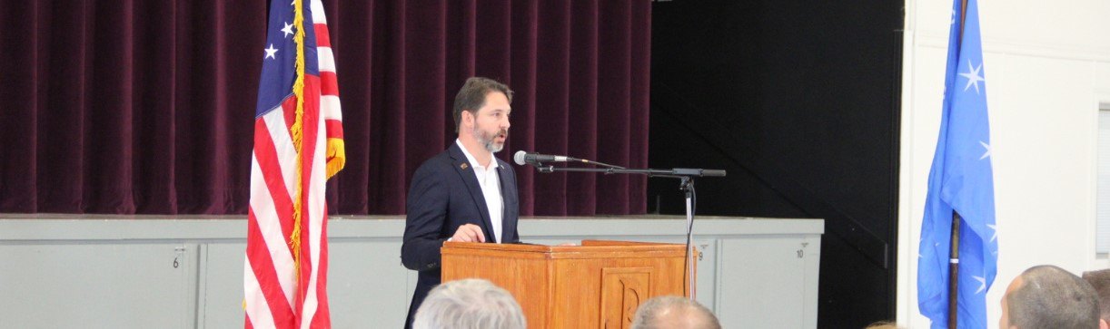 A person in a dark blazer speaks at a wooden podium flanked by American and blue flags.