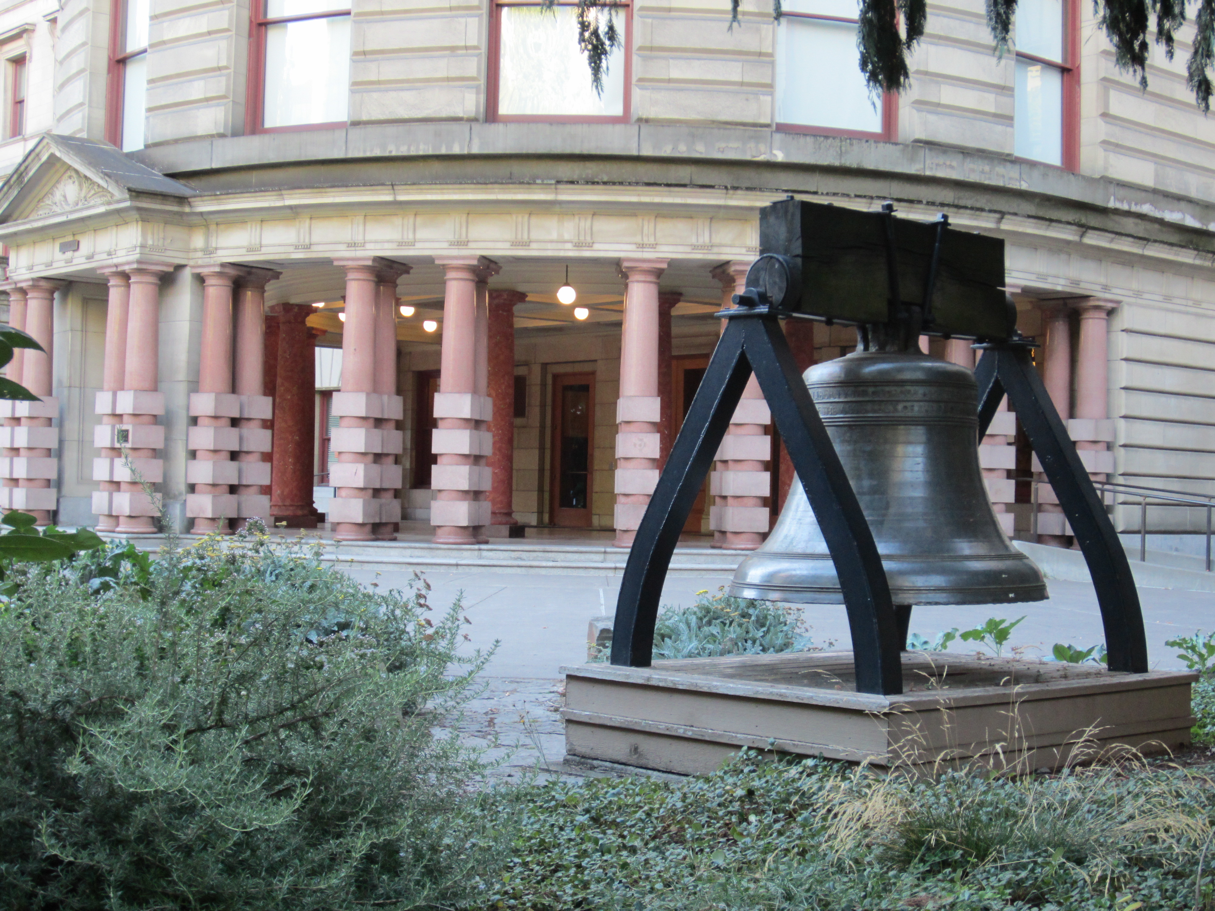 Large bronze bell on a metal stand outside a classical stone government building.