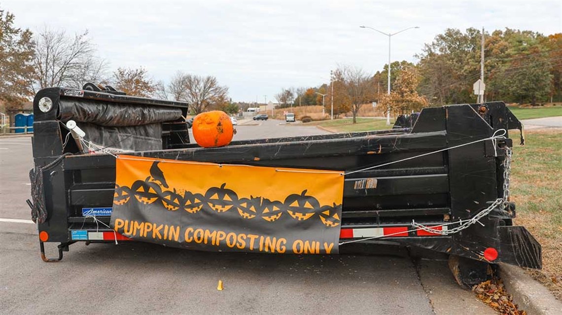 Black dumpster with orange pumpkin composting banner and a pumpkin on top, parked in a lot with autumn trees.