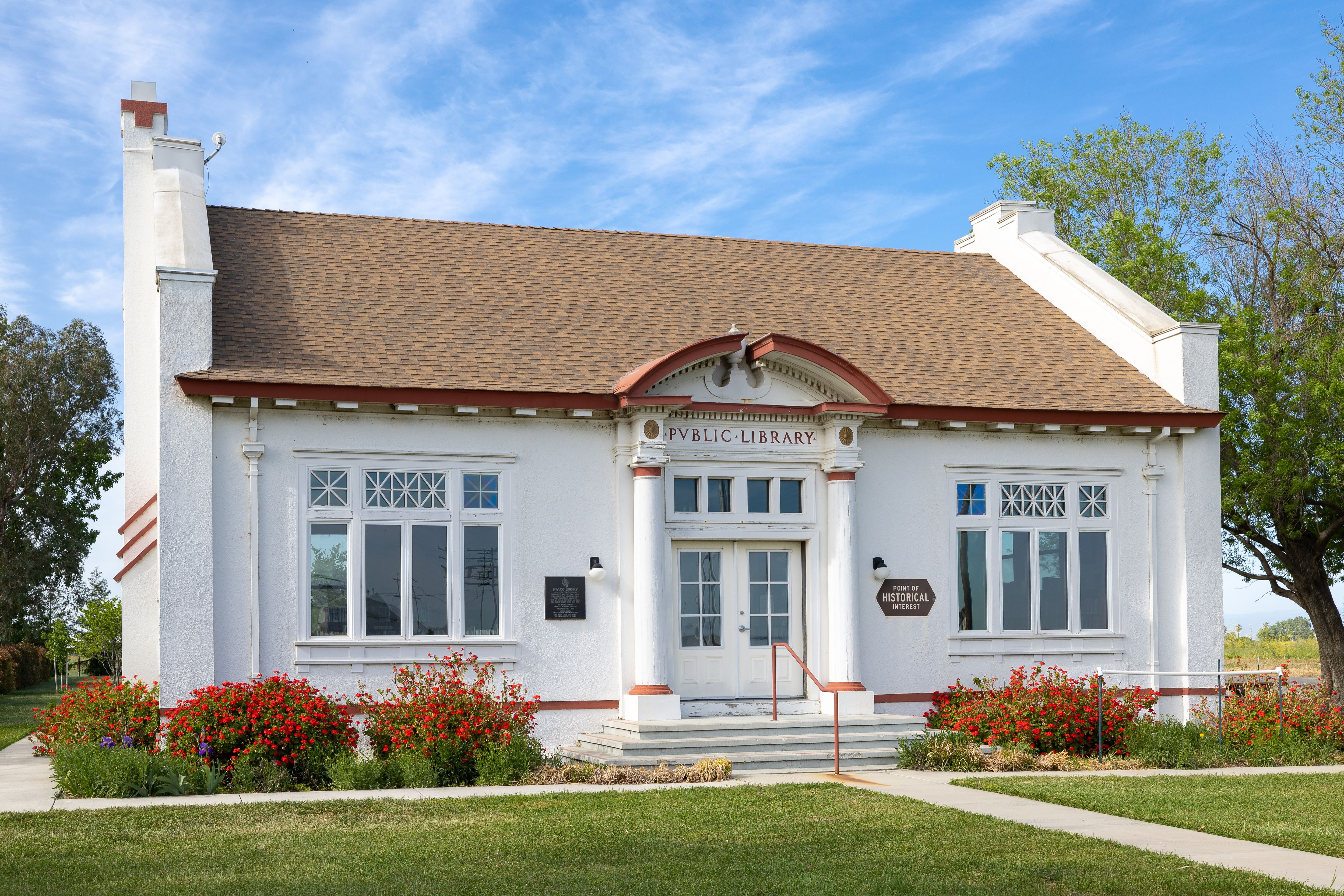 Small white Classical Revival public library building with columns and flowering shrubs.