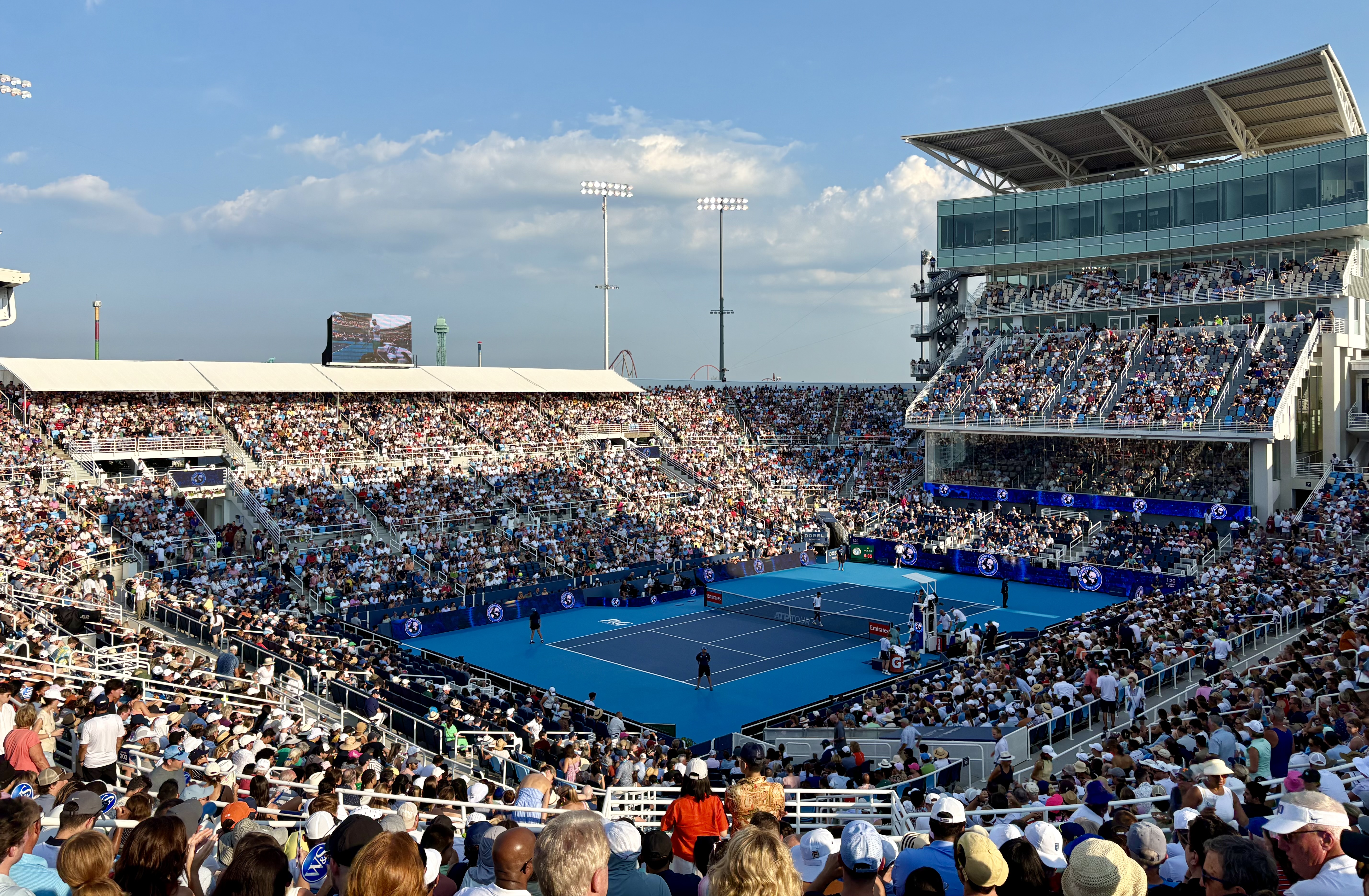 Packed outdoor tennis stadium with a blue hard court and full grandstands under a partly cloudy sky.