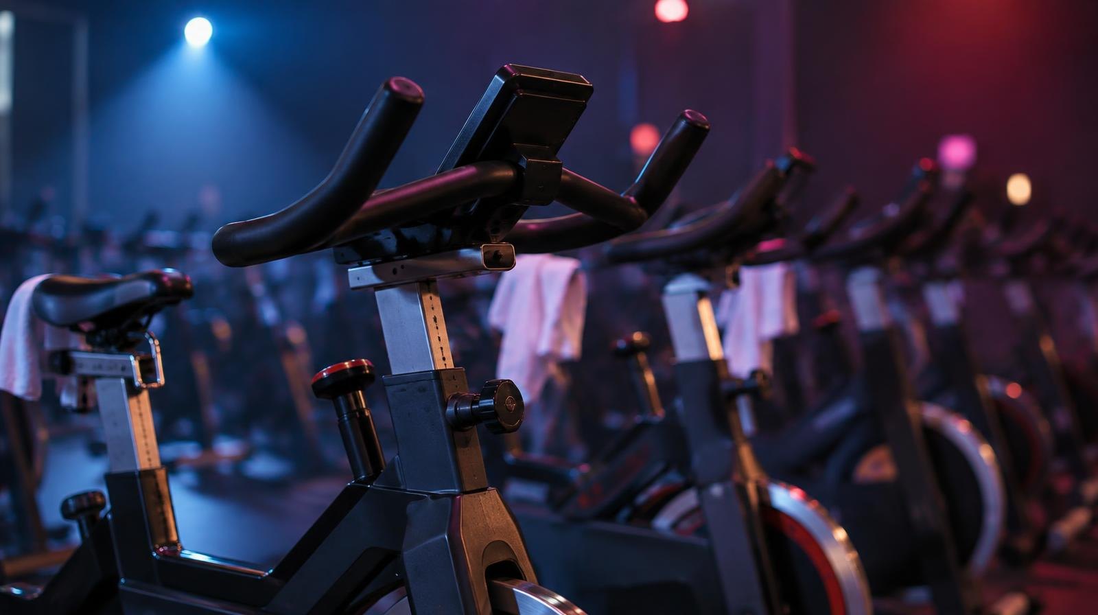Row of stationary spin bikes in a darkened studio lit by colorful stage lighting.