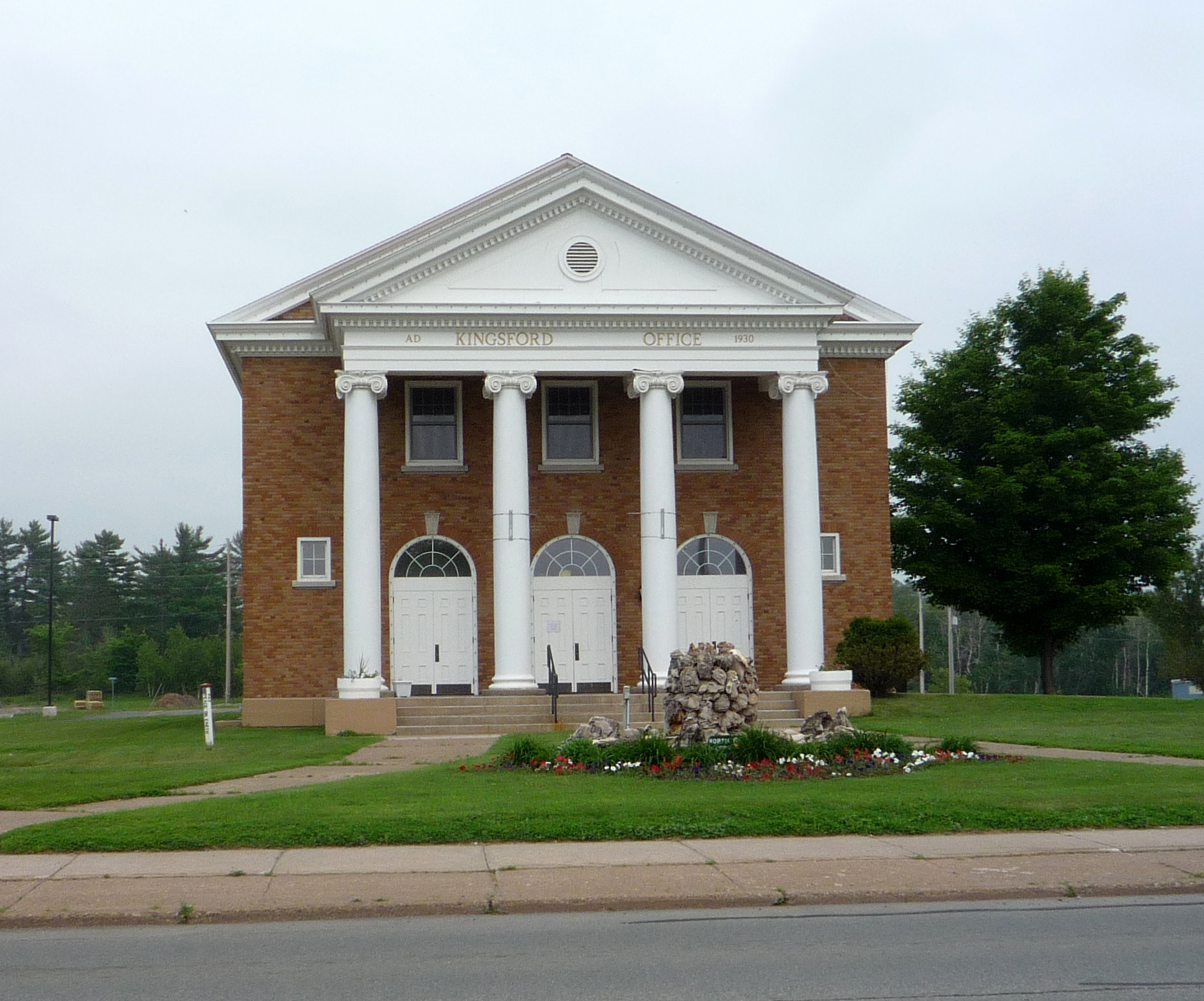 Red brick city hall with white columns and arched doors on a landscaped roundabout.