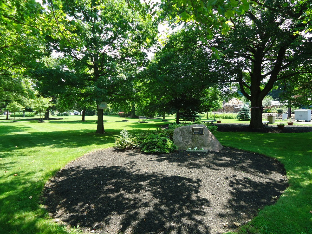 Landscaped public park with mature shade trees and a boulder monument on a green lawn.