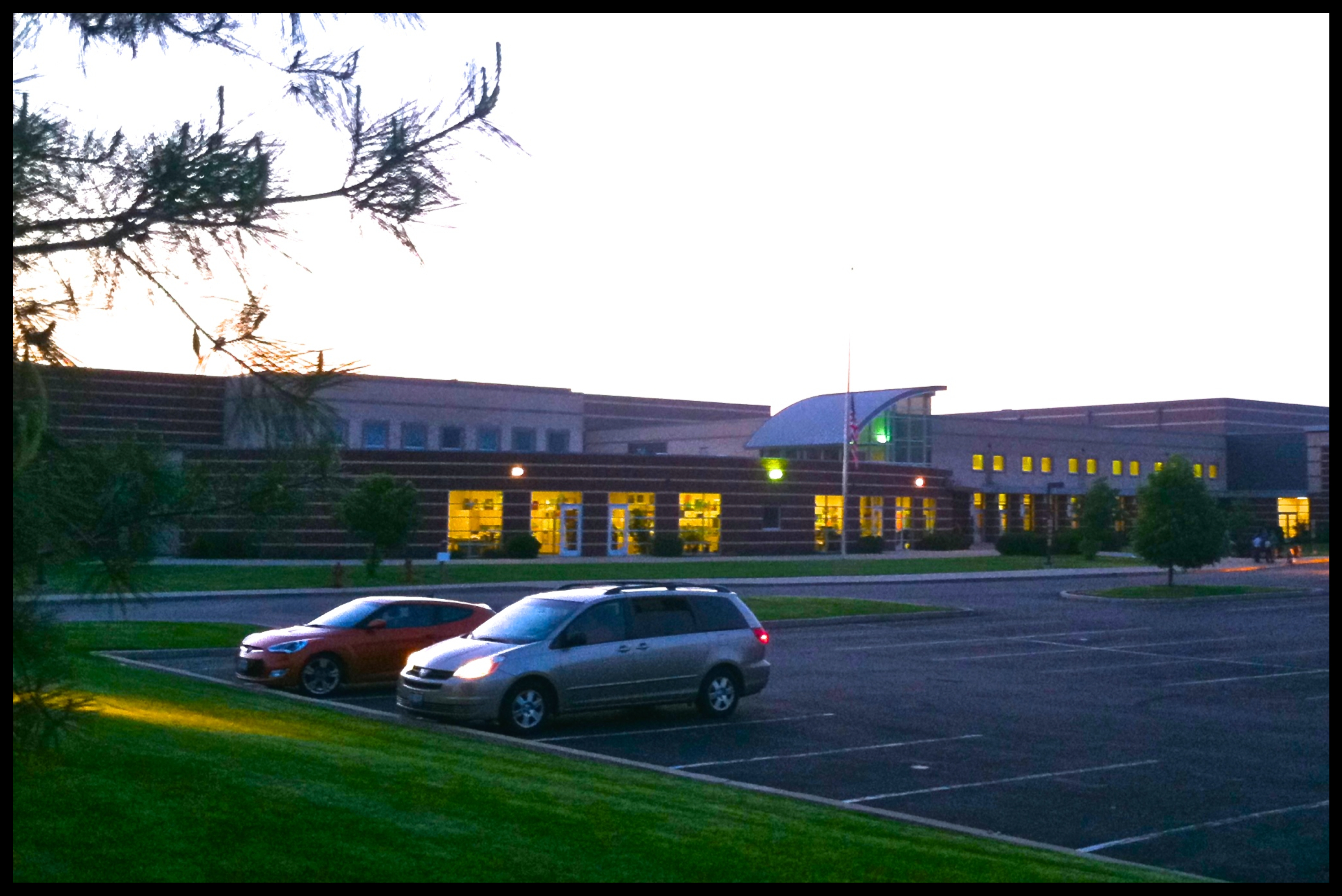 Large brick high school building at dusk with interior lights on and a parking lot in front.