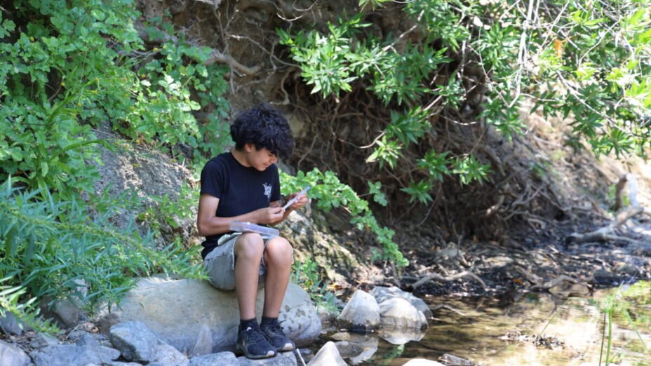 A child sits on a rock beside a shallow, rocky creek, examining something in his hands.