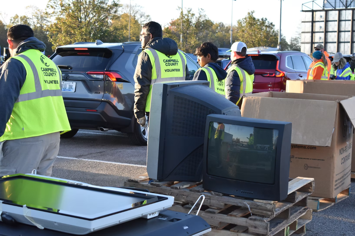 Volunteers in yellow vests stack old CRT televisions on a pallet at an outdoor drop-off event.