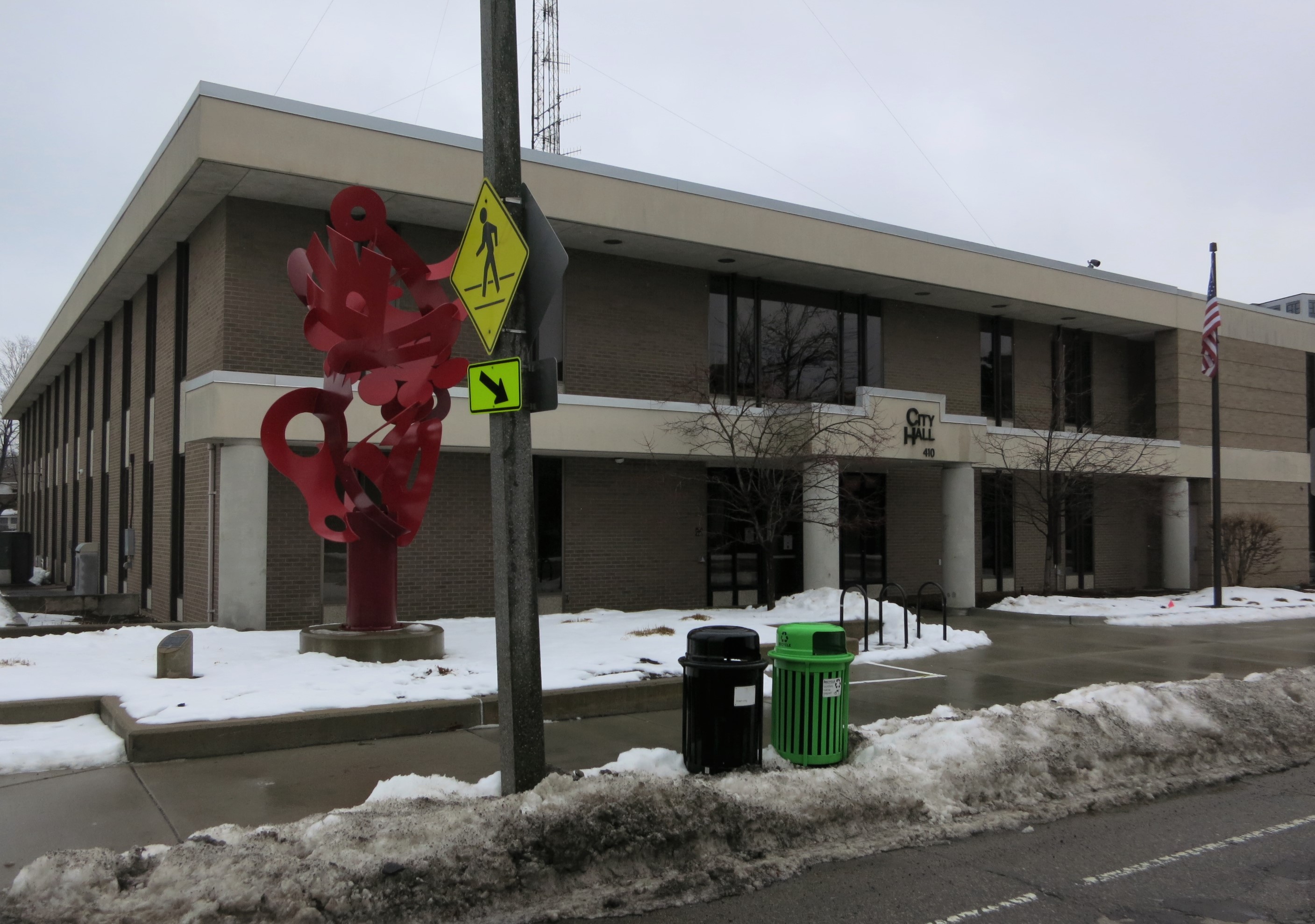 Two-story brick city hall building with a red sculpture and American flag in winter.