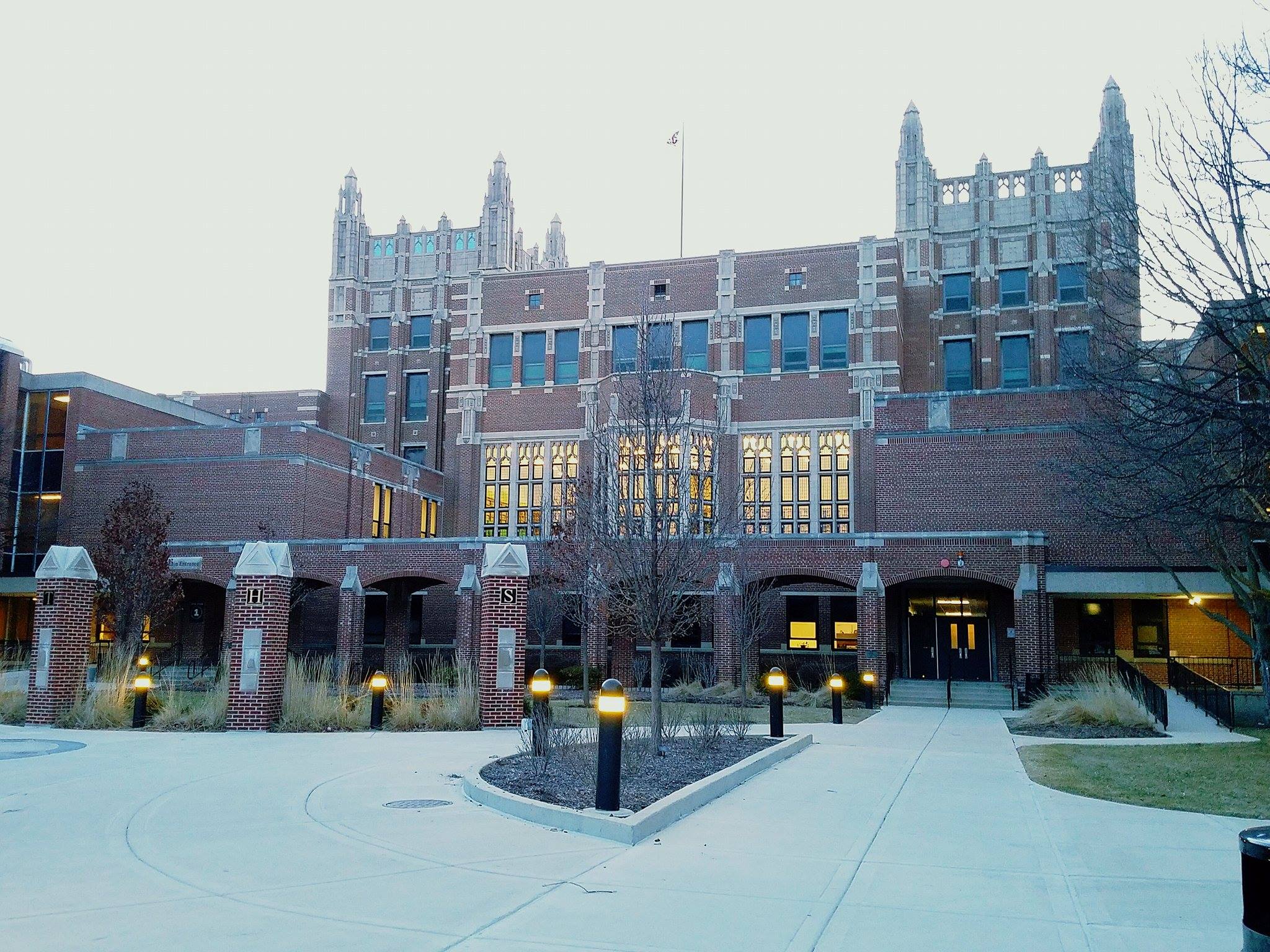 Red-brick Gothic-style high school building at dusk, with pathway lights and bare trees.