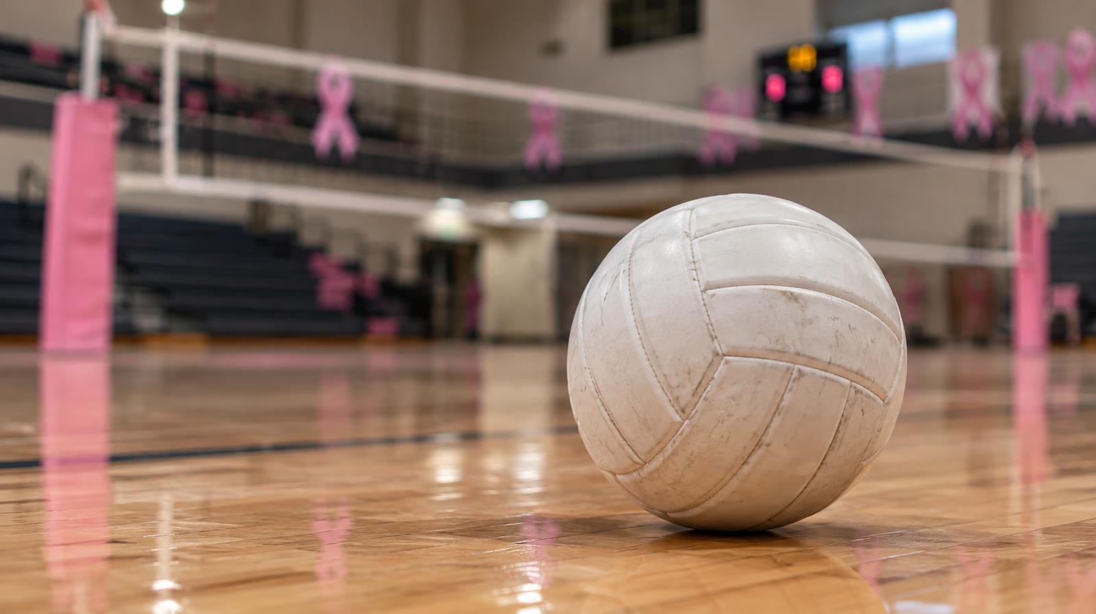 A white volleyball on a hardwood gymnasium floor near the net with pink decorations in the background.