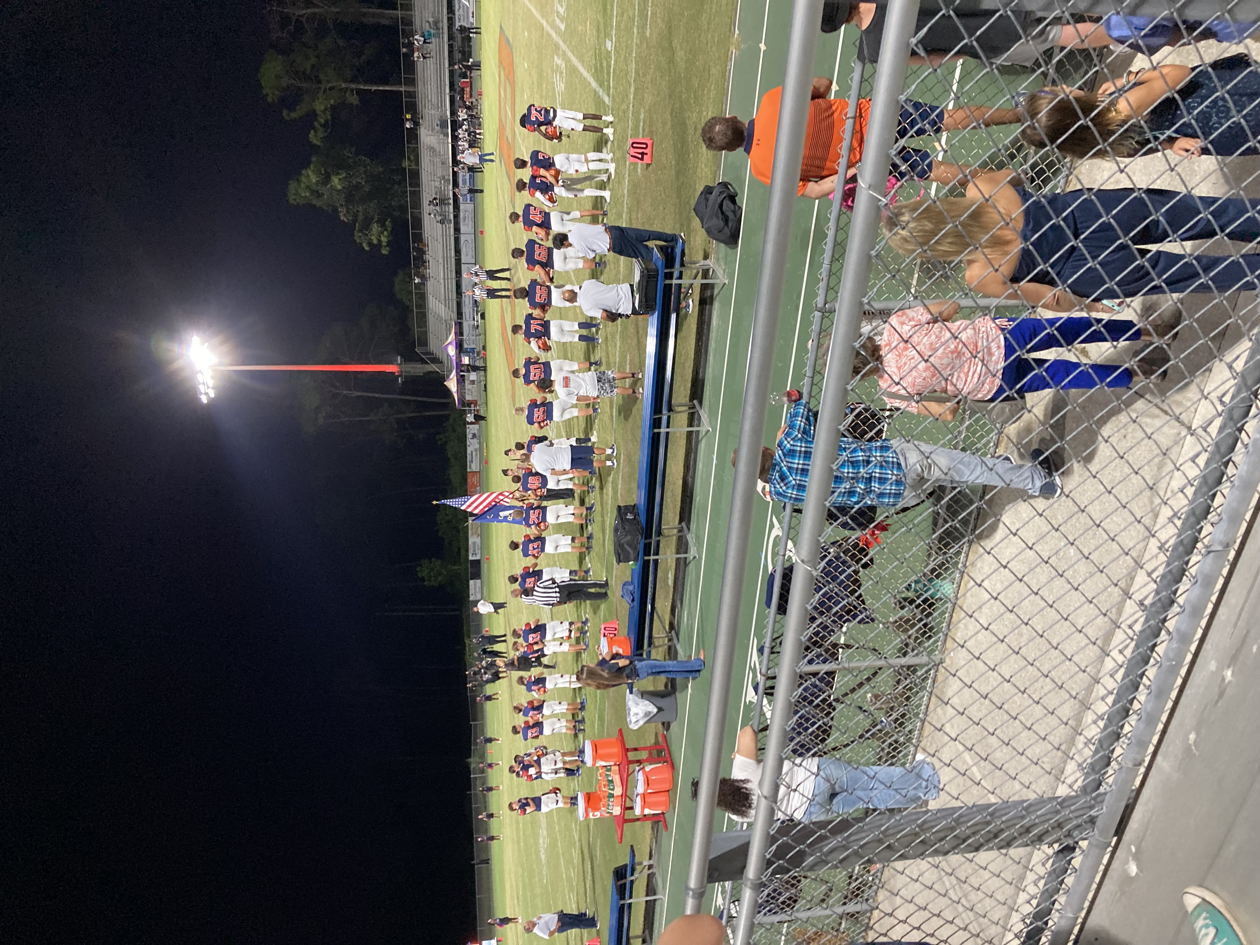 Football players standing on a sideline at night under bright stadium lights.