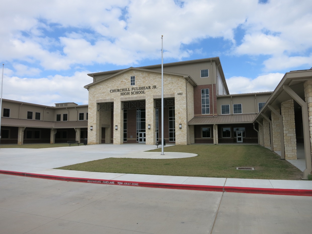Front entrance of a limestone-clad high school building with a flagpole and green lawn.