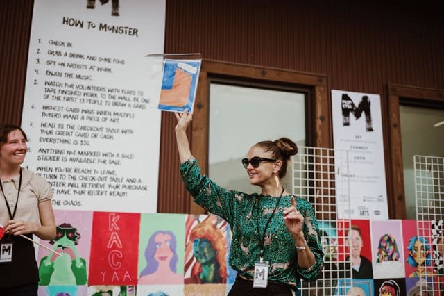 A woman holds up a small painting at an outdoor art event with colorful works displayed on a fence behind her.