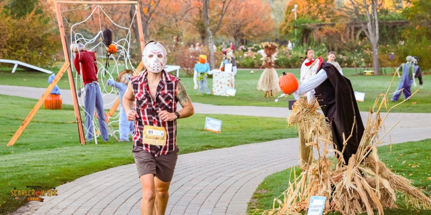 A masked runner in race bib jogs a brick garden path lined with scarecrows and hay bales.