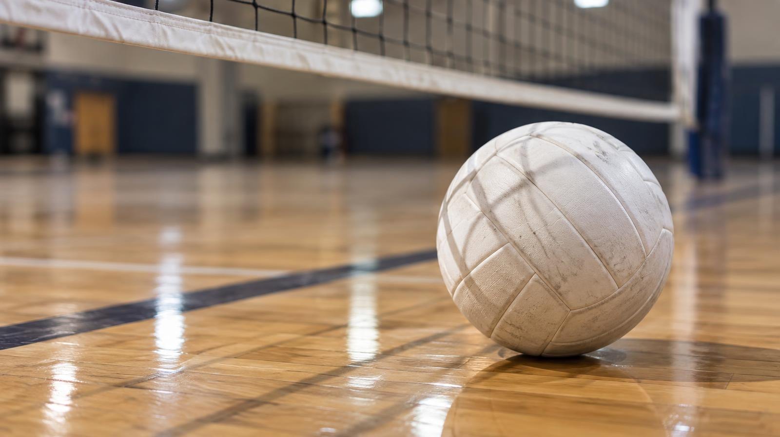 A white volleyball resting on a hardwood gym floor near a net.