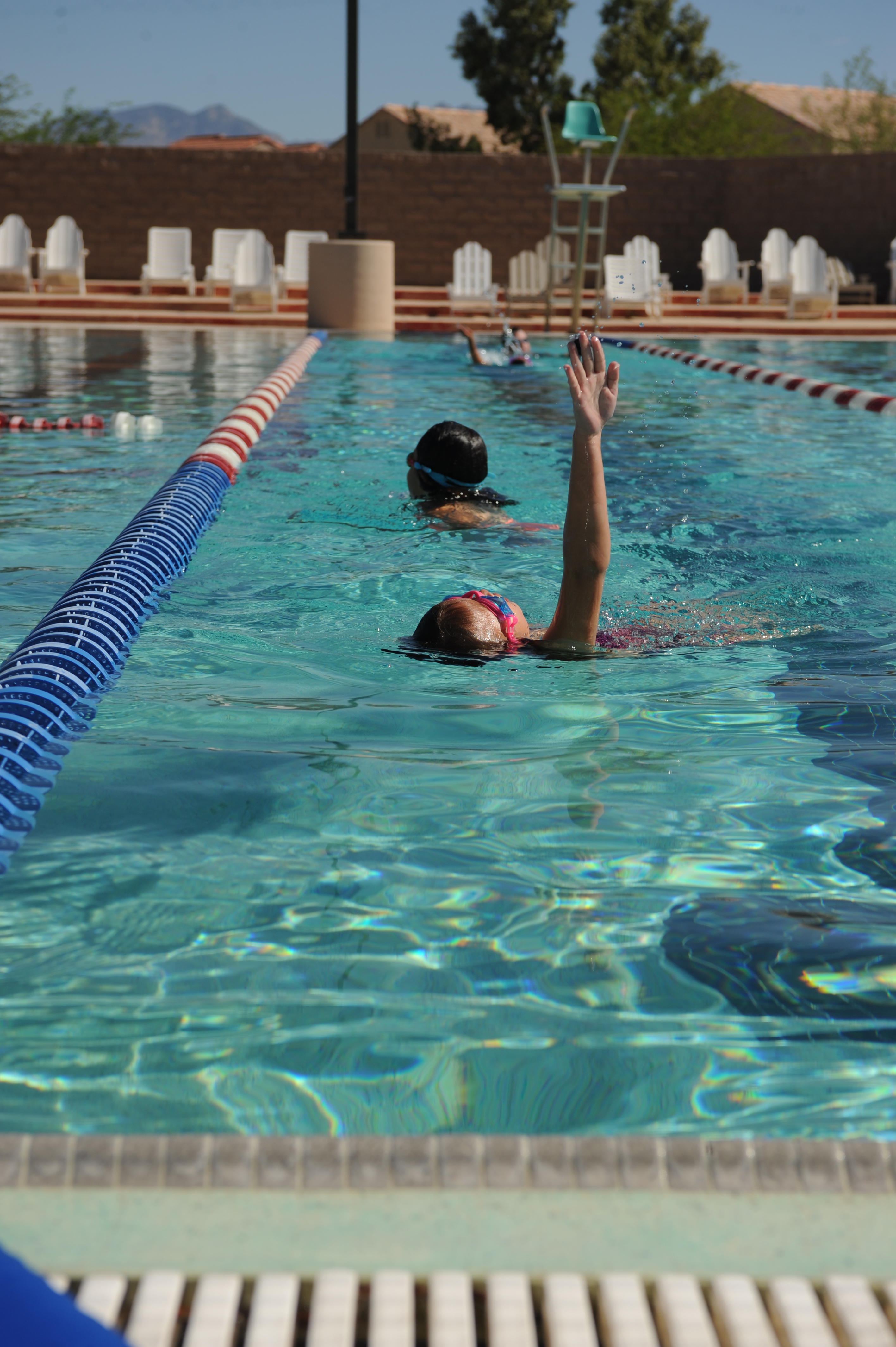 Two swimmers in an outdoor lap pool with red-and-white lane dividers, one mid-backstroke.