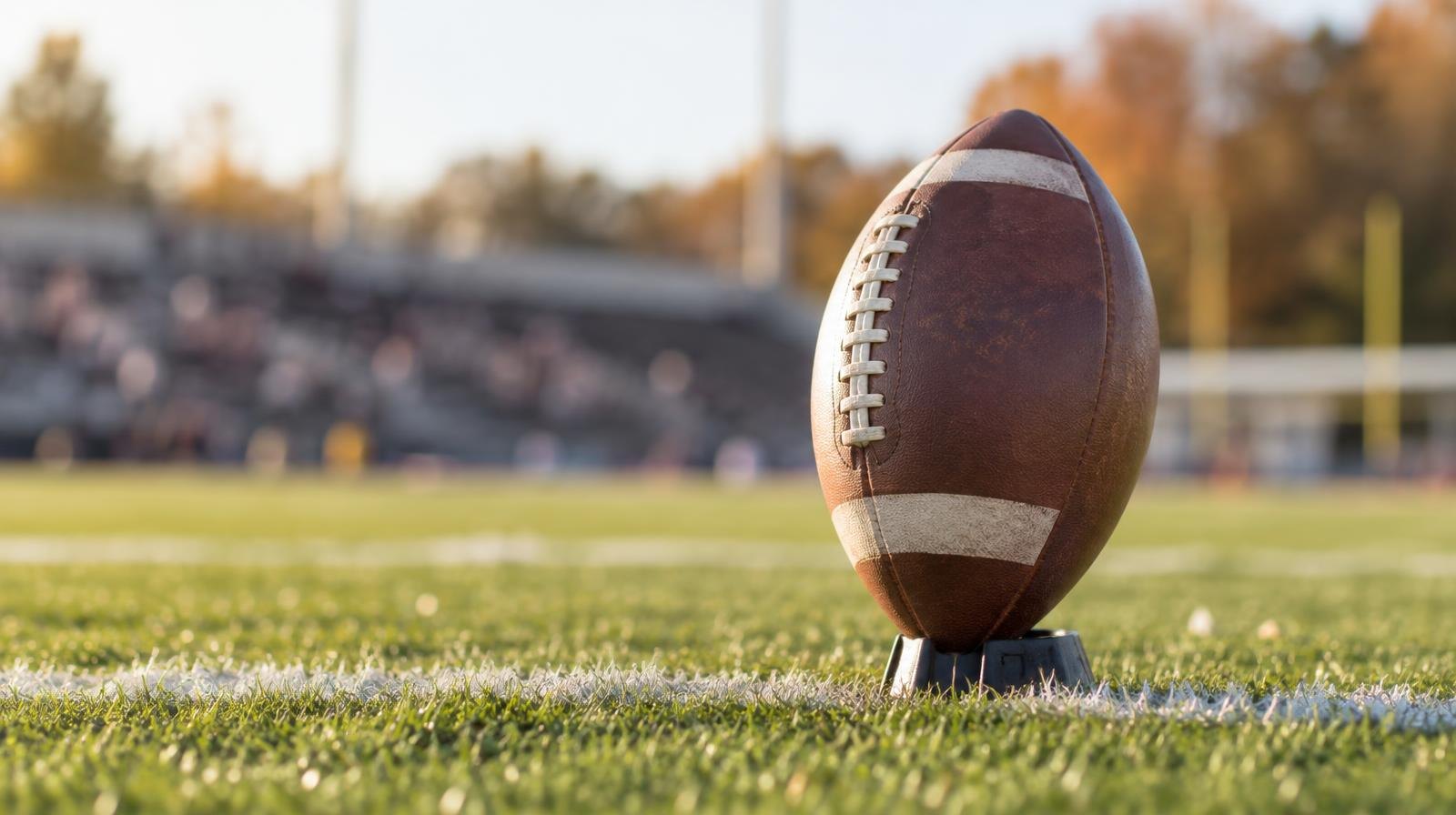 A football on a tee positioned on a grass field with white yard-line markings.