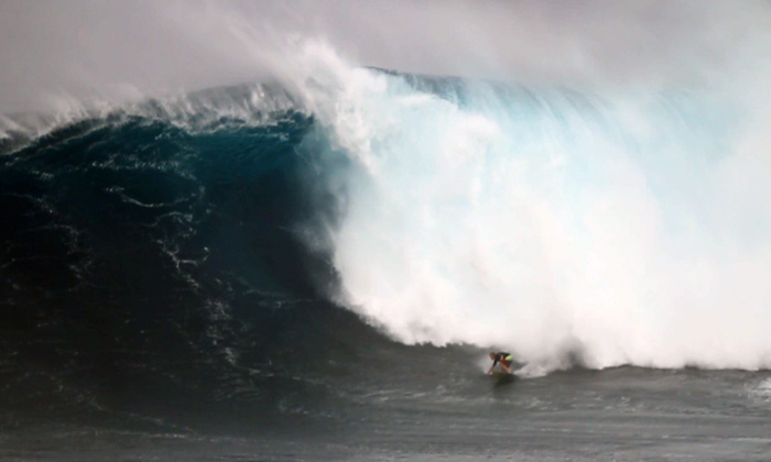 A lone surfer rides at the base of a massive, towering breaking wave.