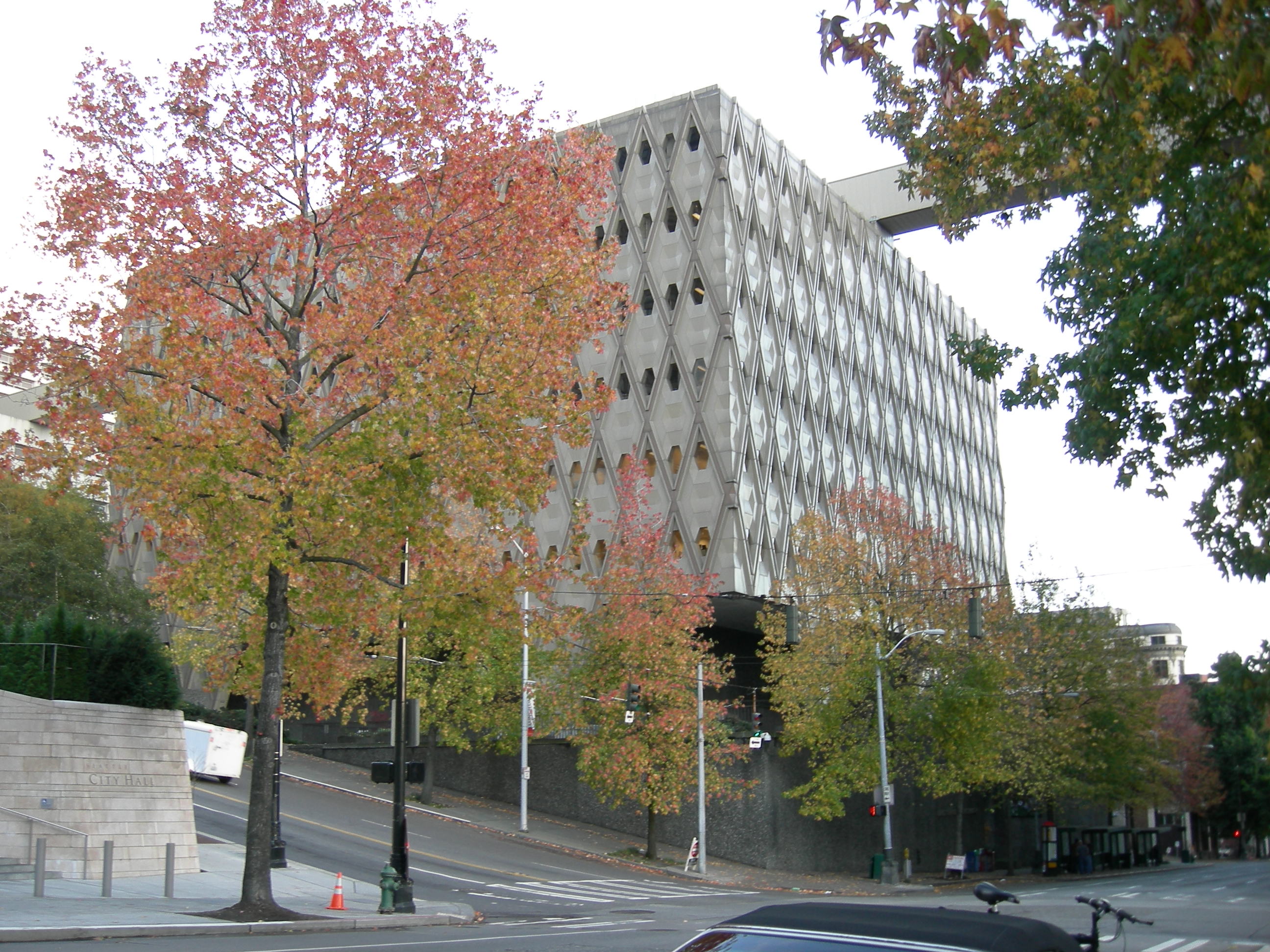 Multi-story modernist government building with diamond-patterned concrete facade and autumn trees along the street.