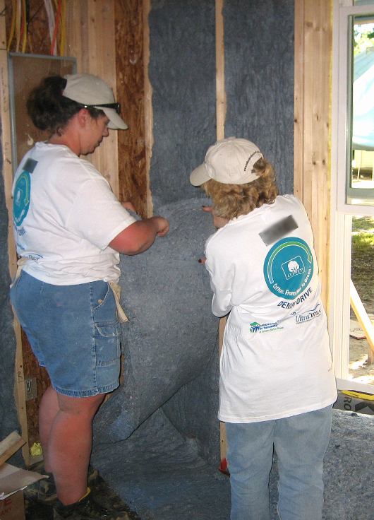Two volunteers in Habitat for Humanity shirts install insulation in a framed house.