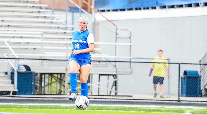 Female soccer player in blue uniform number 19 dribbling a ball on a turf field.