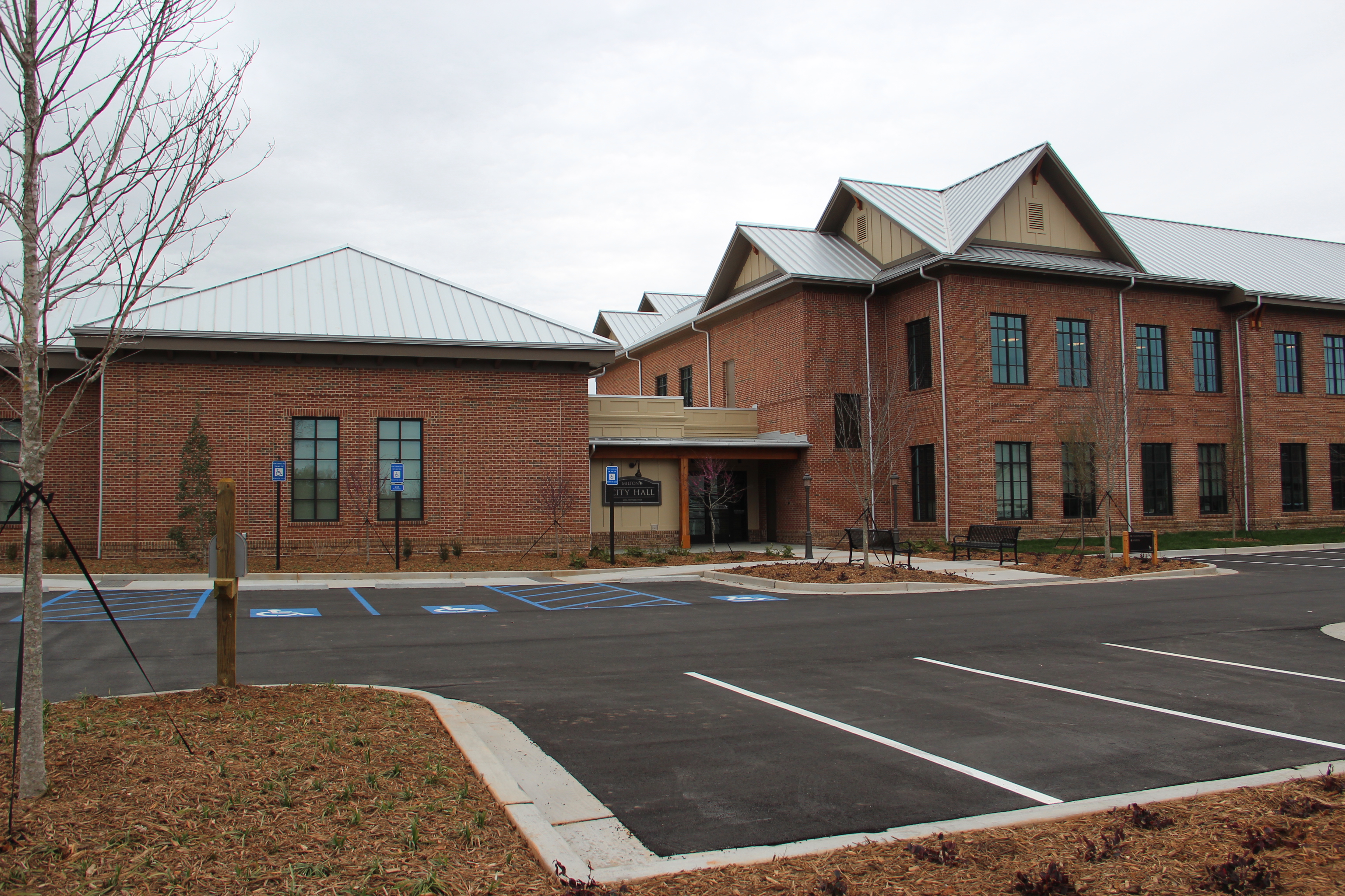 Two-story red brick municipal building with metal roof and bare trees under an overcast sky.