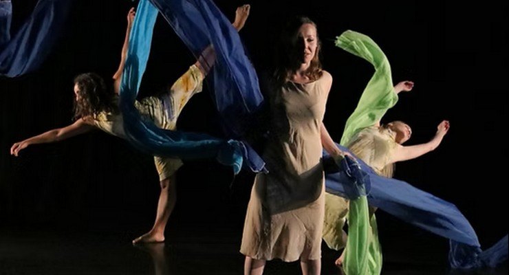 Dancers in neutral dresses swirl blue, green, and yellow fabric on a dark stage.