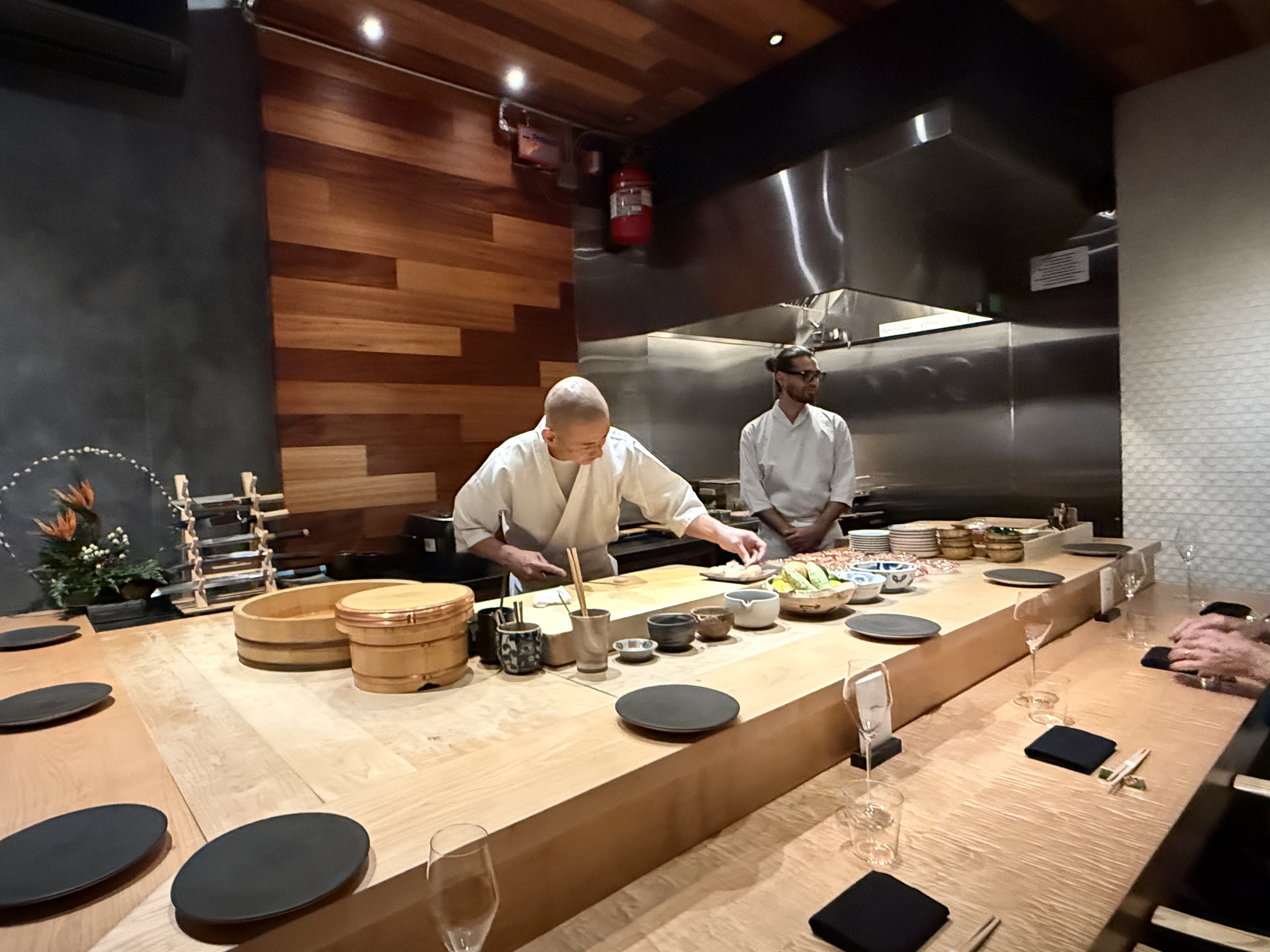 Two chefs in white jackets working behind a wooden sushi counter.