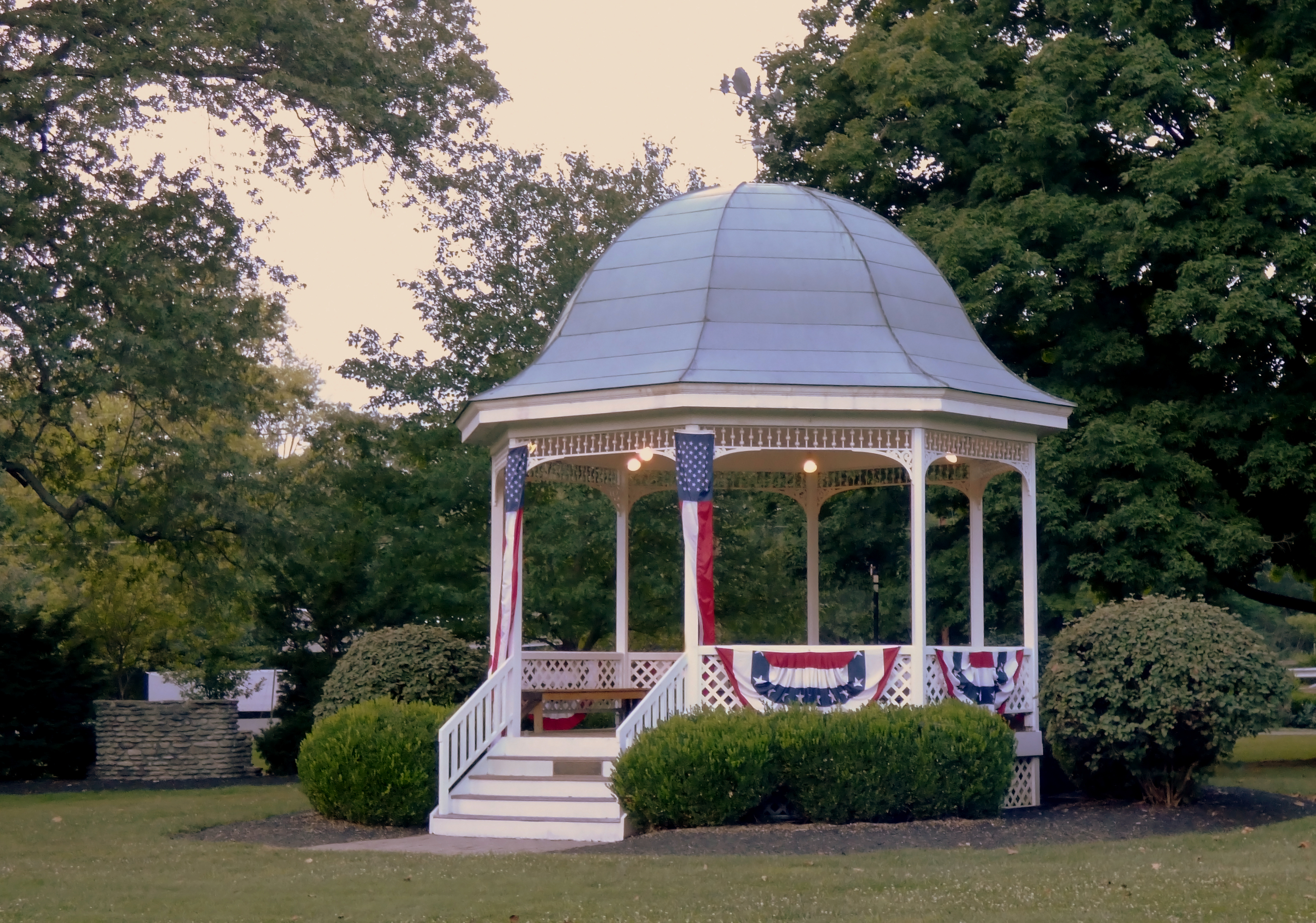 White gazebo with domed roof and patriotic bunting, surrounded by green shrubs and trees.