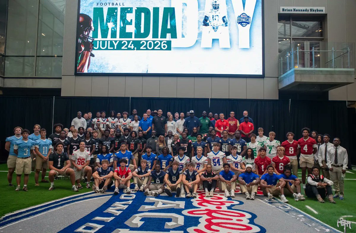 Large group of football players and coaches posing indoors at a media day event.