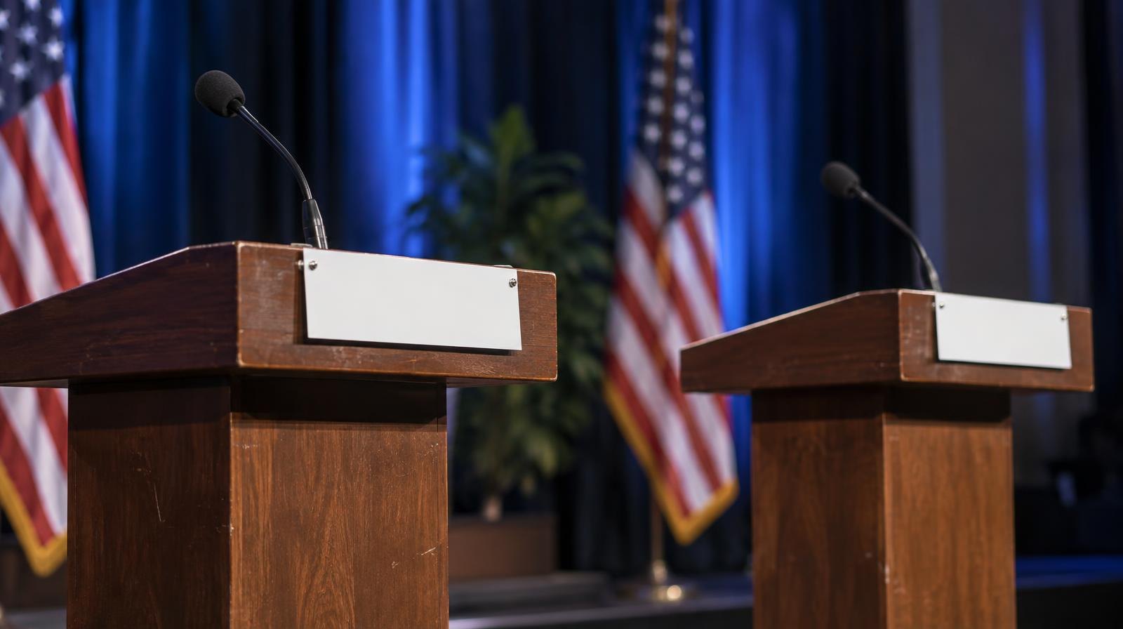 Two empty podiums with microphones face each other on a brightly lit stage.