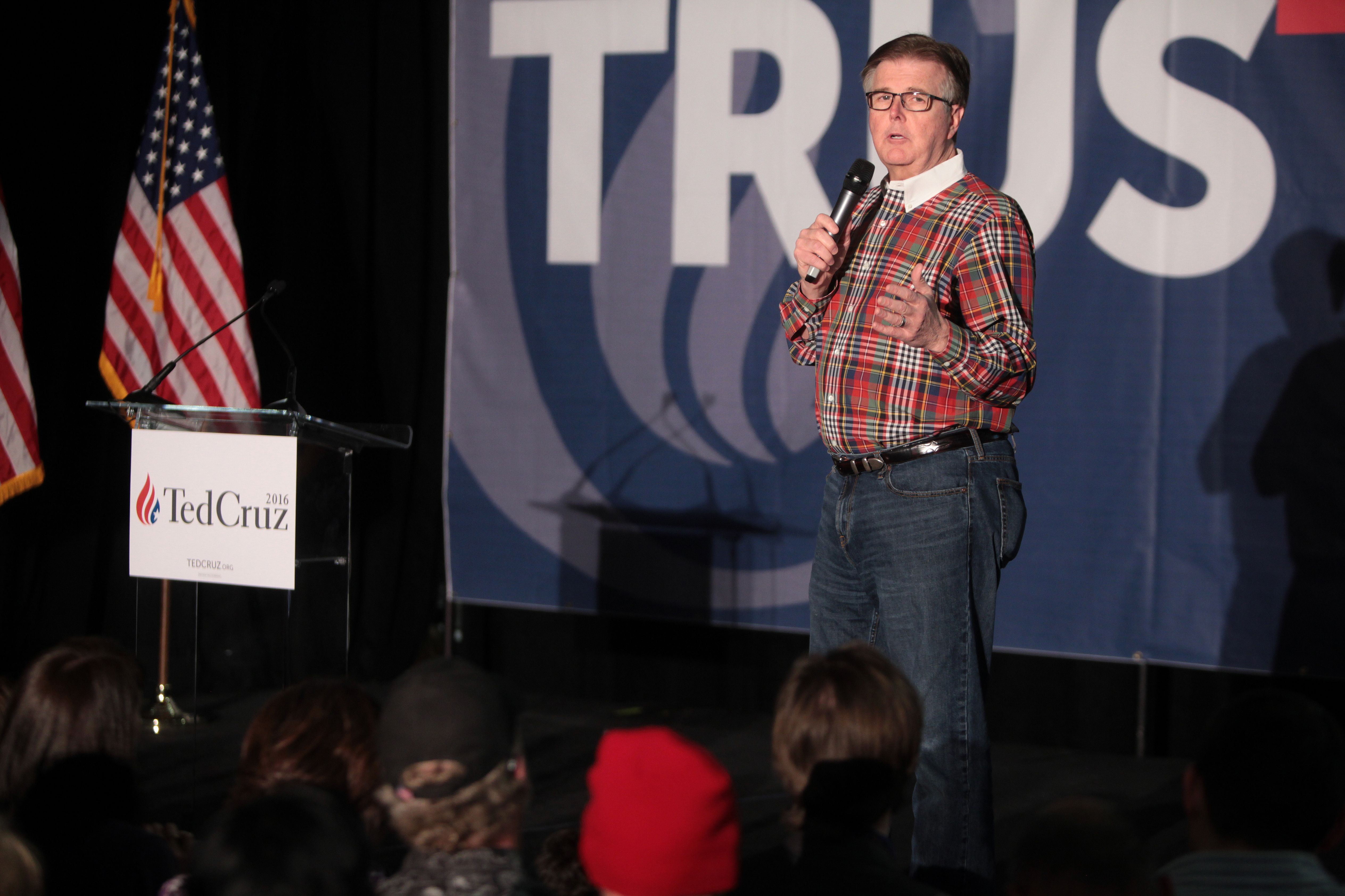 A man in a plaid shirt speaks into a microphone at an indoor political event.