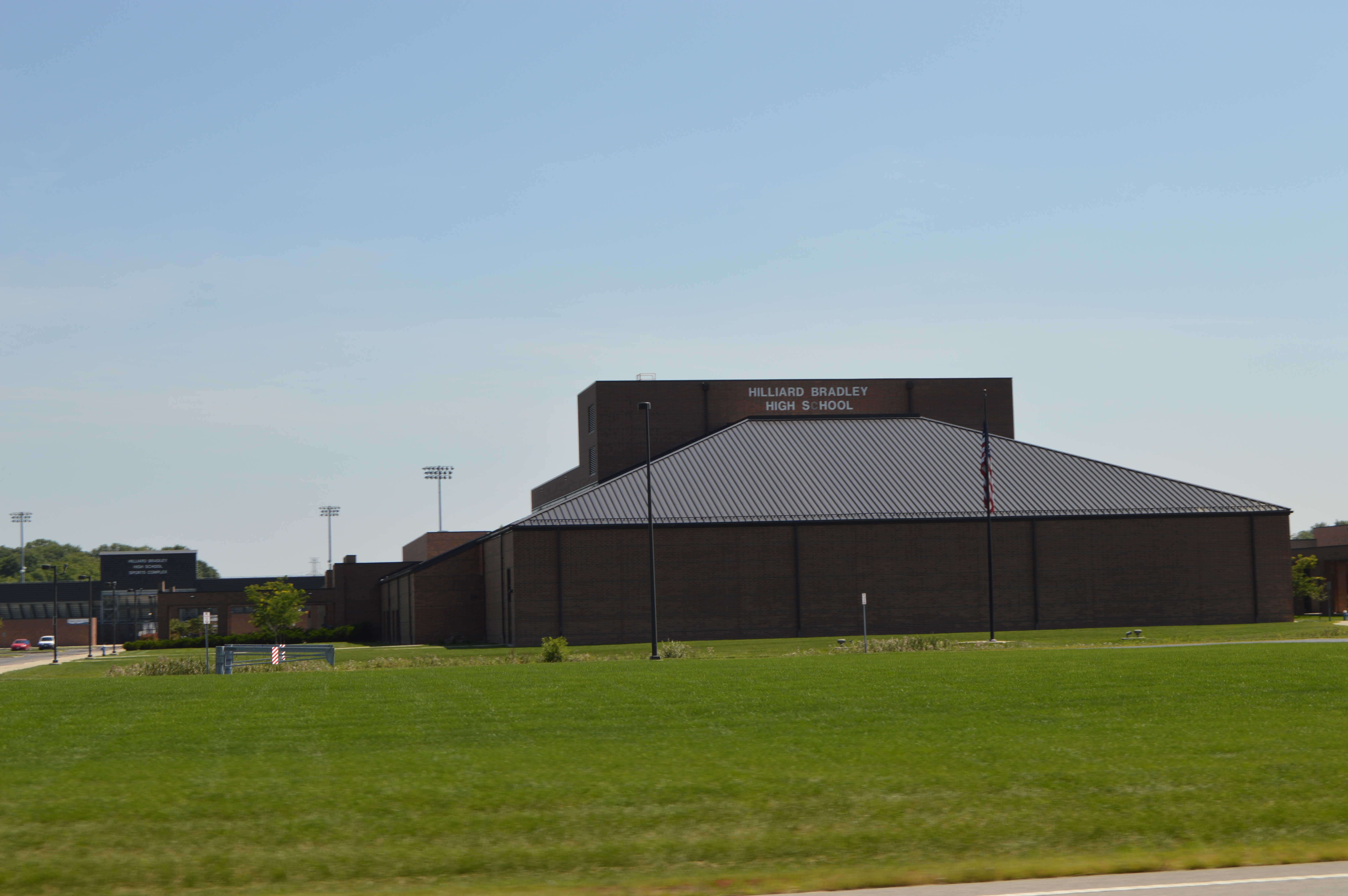 Exterior of a brick high school building with a metal roof and stadium lights behind it.