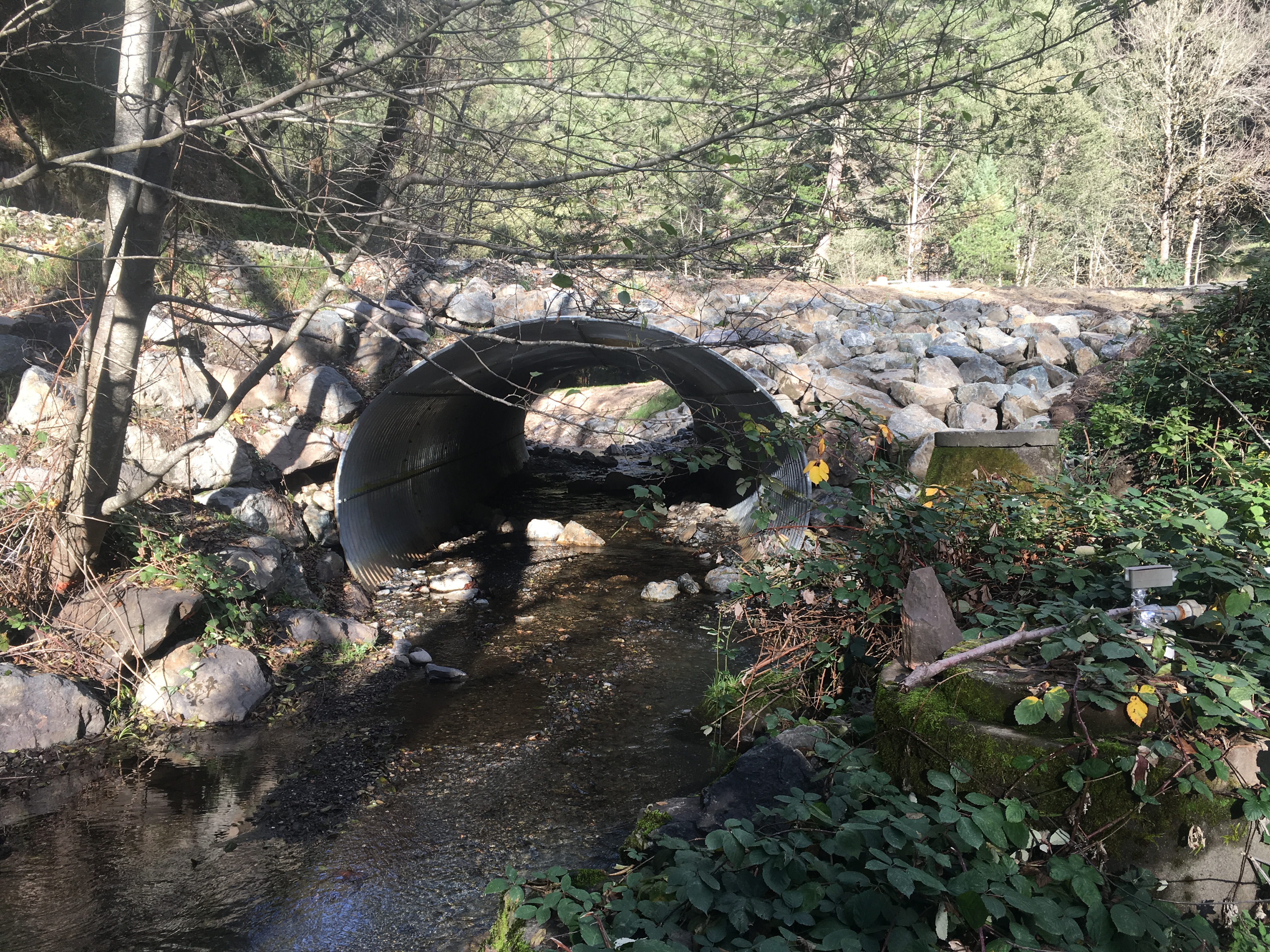 A large metal arch culvert installed beneath a gravel road over a stream channel.
