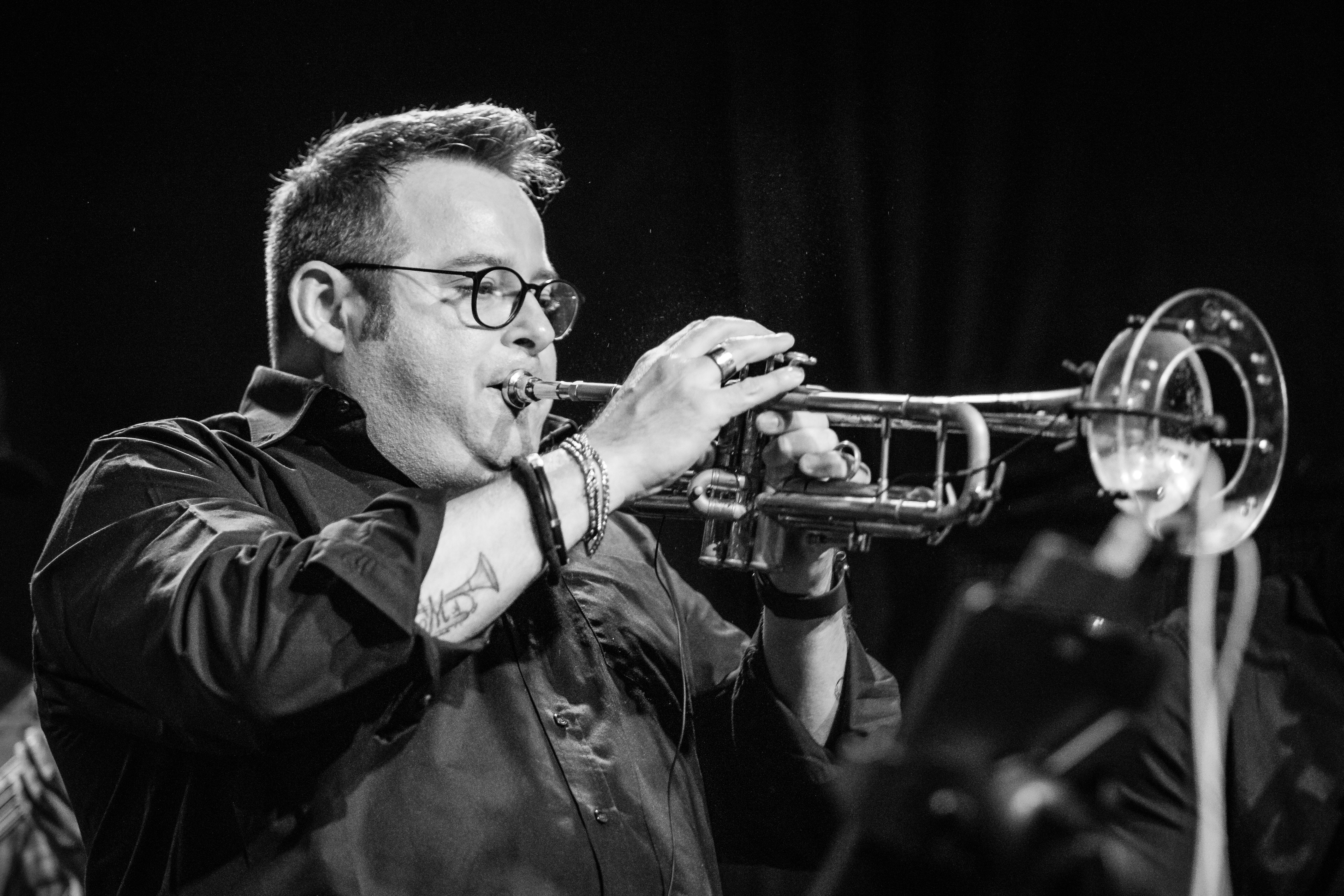 A musician plays trumpet on a darkened stage under a spotlight, in black and white.