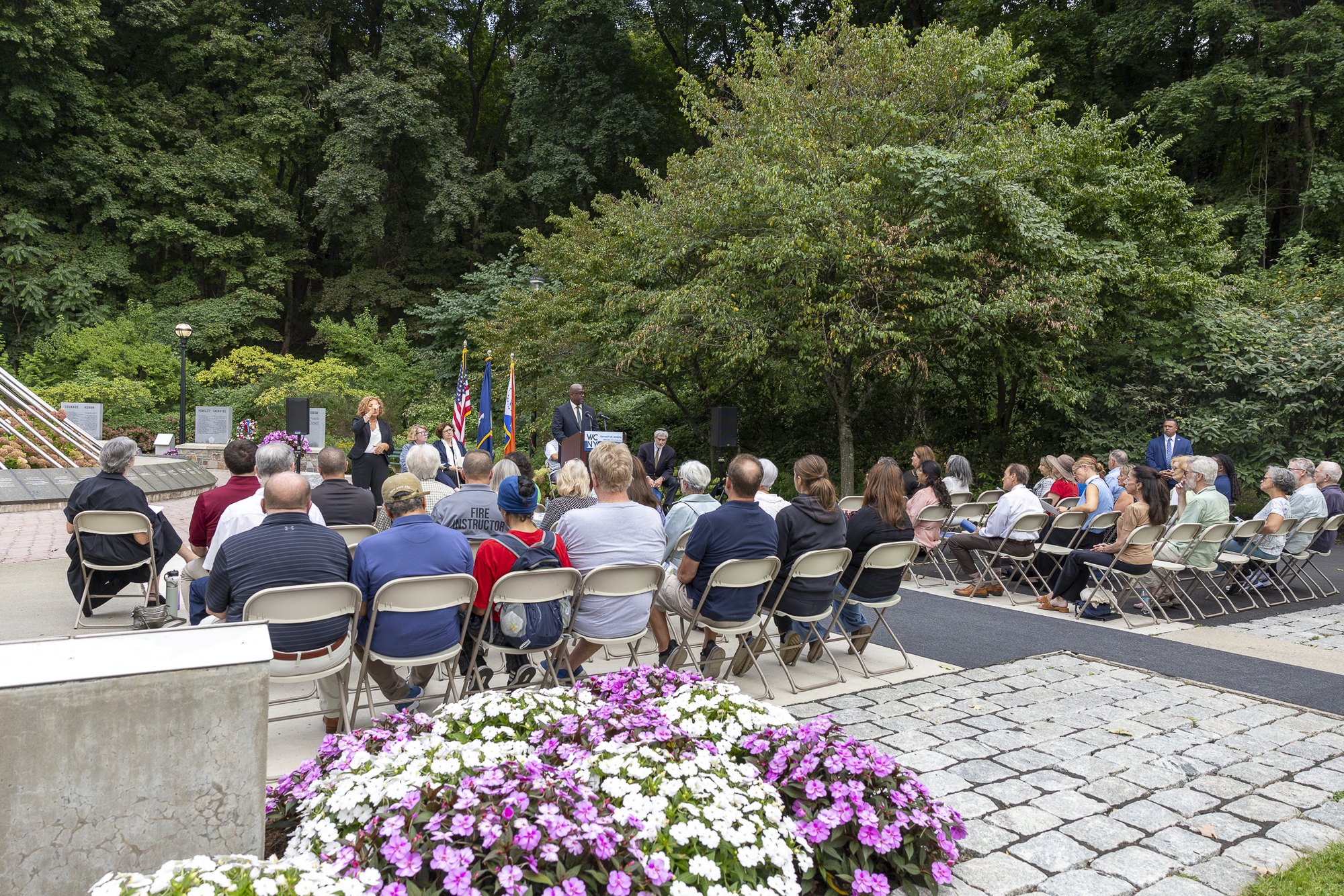 Crowd seated in folding chairs at an outdoor ceremony with flags and a podium.
