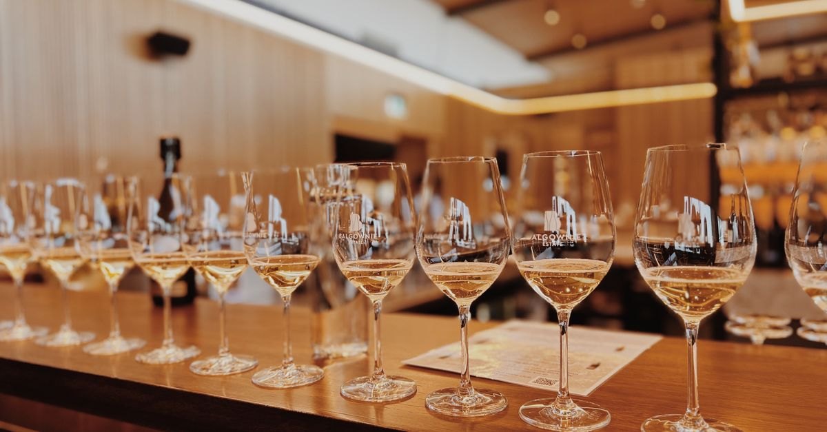 A row of stemmed wine glasses filled with pale golden liquid on a wooden bar counter.