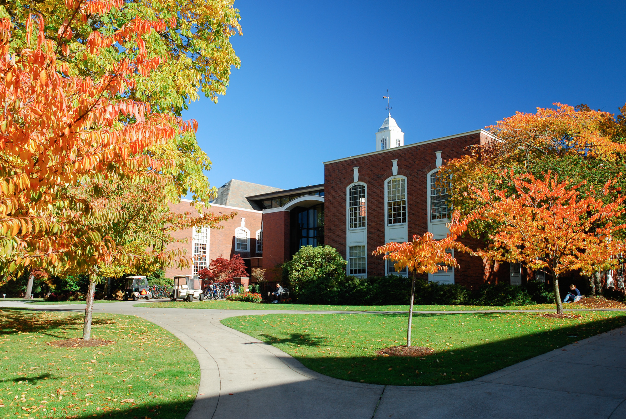 Red-brick university building with white cupola and colorful autumn trees on a curved walkway.