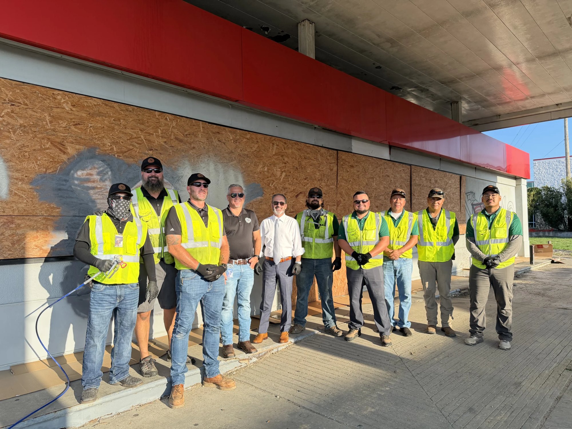 Workers in yellow high-visibility vests stand in front of a boarded-up building with a red facade.
