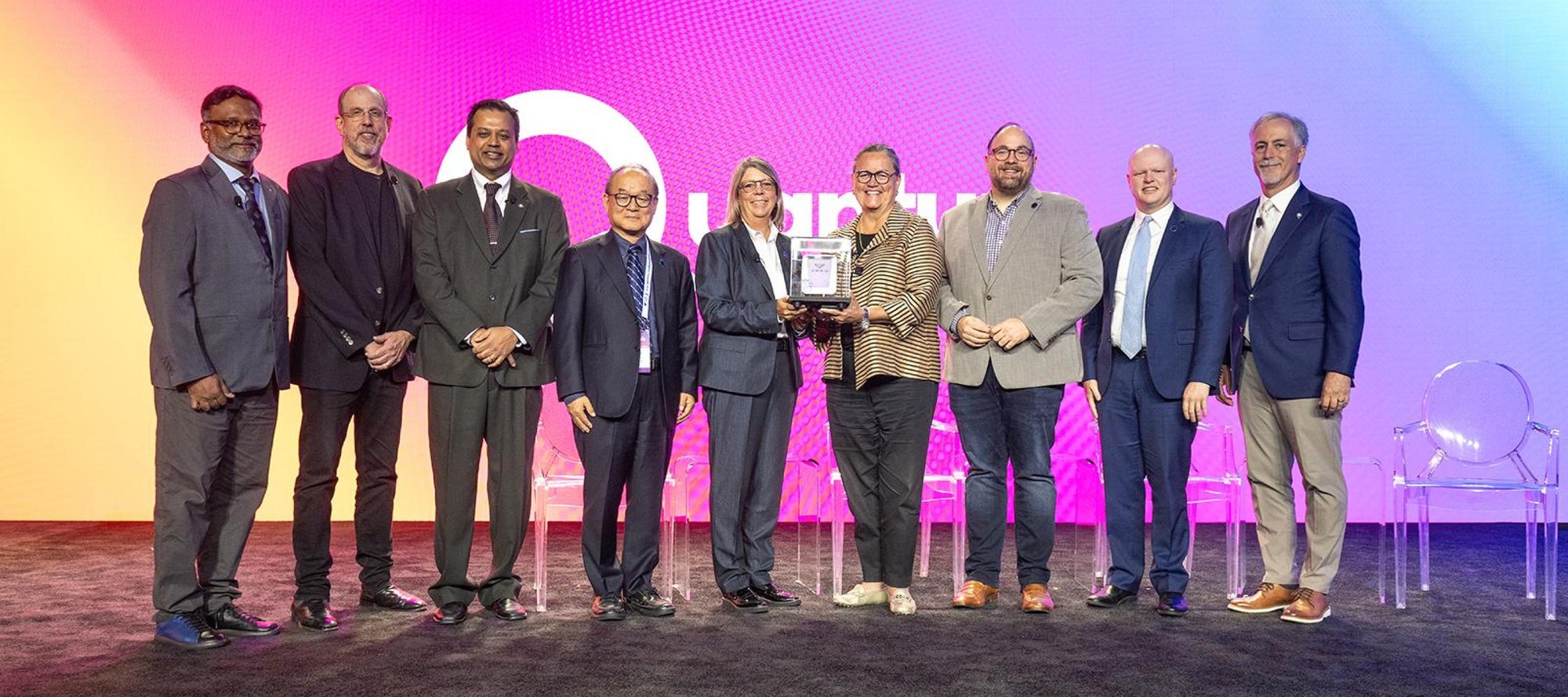 Group of adults in business attire posed on a stage, one holding a framed plaque.