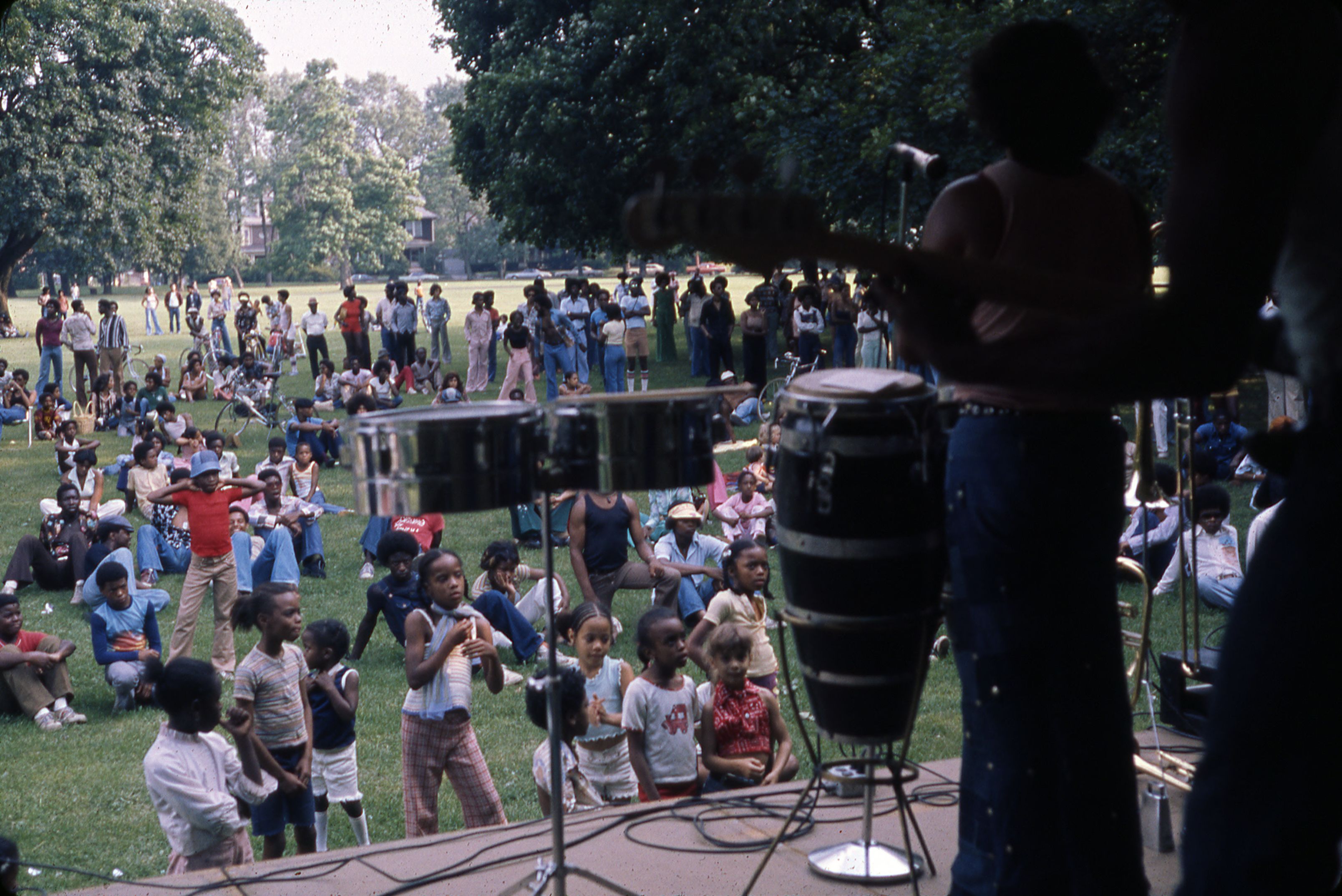 A large crowd sits and stands on a grassy lawn watching an outdoor musical performance.
