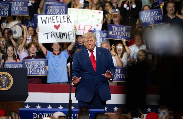 Man in blue suit at podium before large crowd holding signs in a gymnasium.