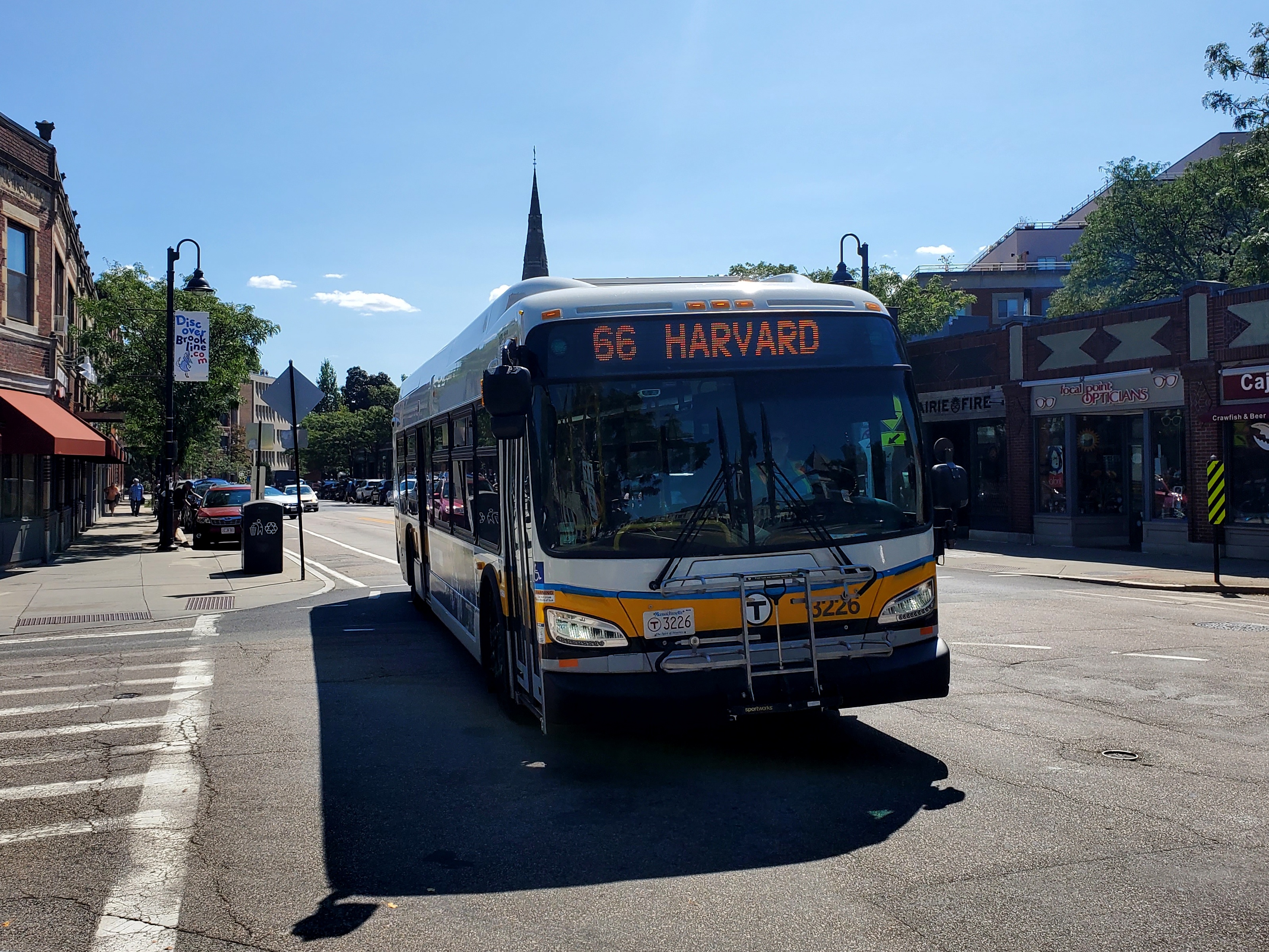 An MBTA bus displaying a Harvard destination sign travels along a commercial street with brick buildings.
