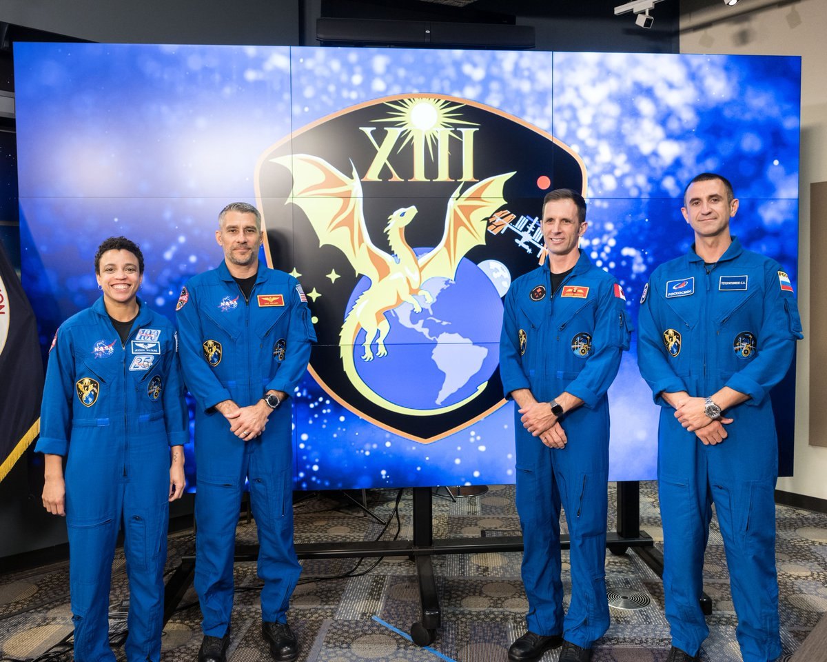 Four people in blue flight suits stand before a large Crew-13 mission patch display.